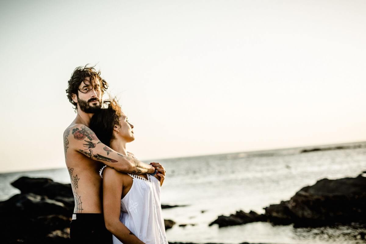 Das Paar steht Arm in Arm am Meer, die Frau guckt in die Sonne, der Mann in die Kamera