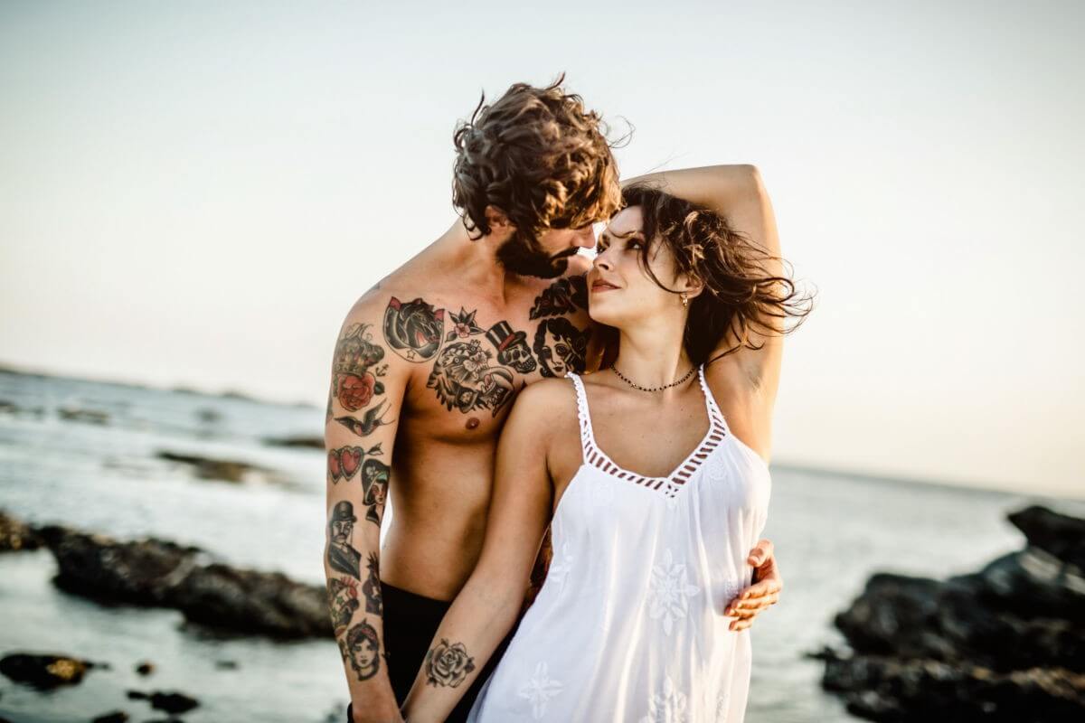 Das Paar steht am Strans mit dem Meer im Hintergrund, der Wind zerzaust die Haare, sie sehen sich in die Augen